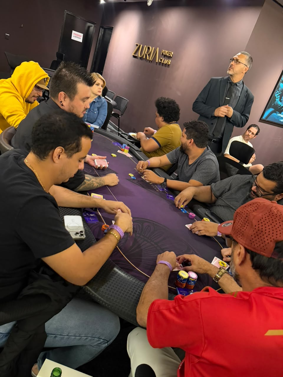 Salao do Zuria Poker Club numa noite de torneio: mesa de poker completa com jogadores e o letreiro dourado Zuria Poker Club na parede