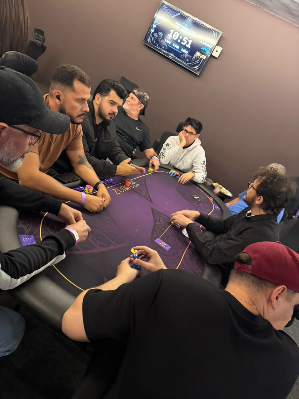 Mesa de poker cheia com a televisao do relogio de torneio na parede mostrando o nivel e os blinds