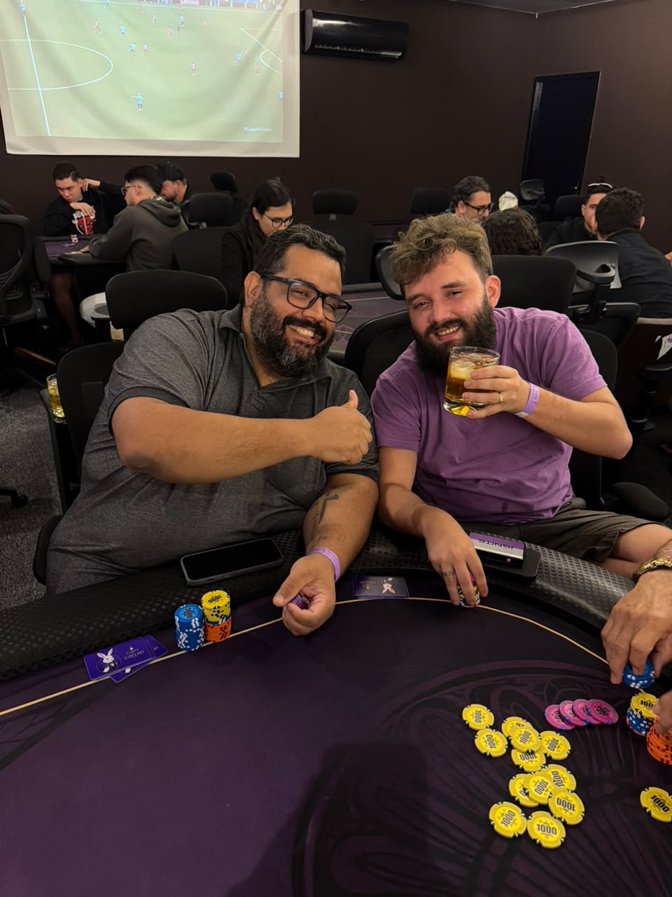 Dois jogadores sorrindo a mesa de poker, com o telao do salao ao fundo