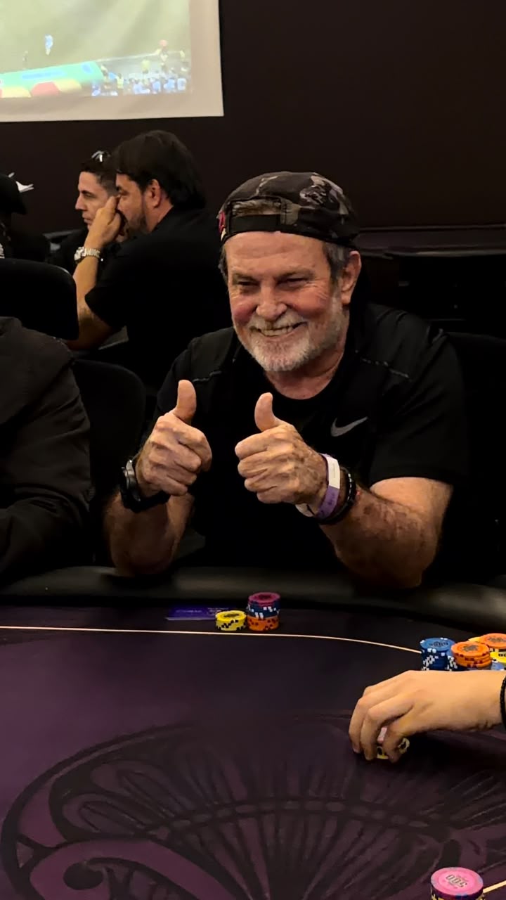 Jogador mais velho sorrindo e fazendo sinal de positivo com as duas maos na mesa de poker